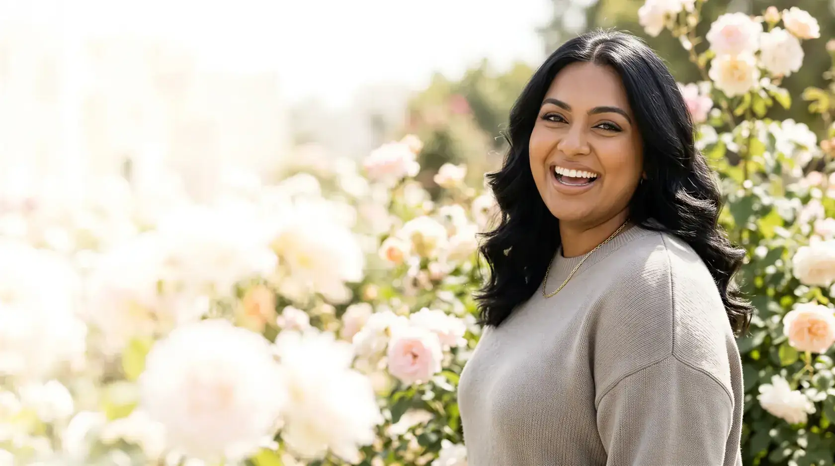 Woman with dark wavy hair smiling in a sunlit Dallas rose garden, reflecting the longer-lasting, natural results of Daxxify