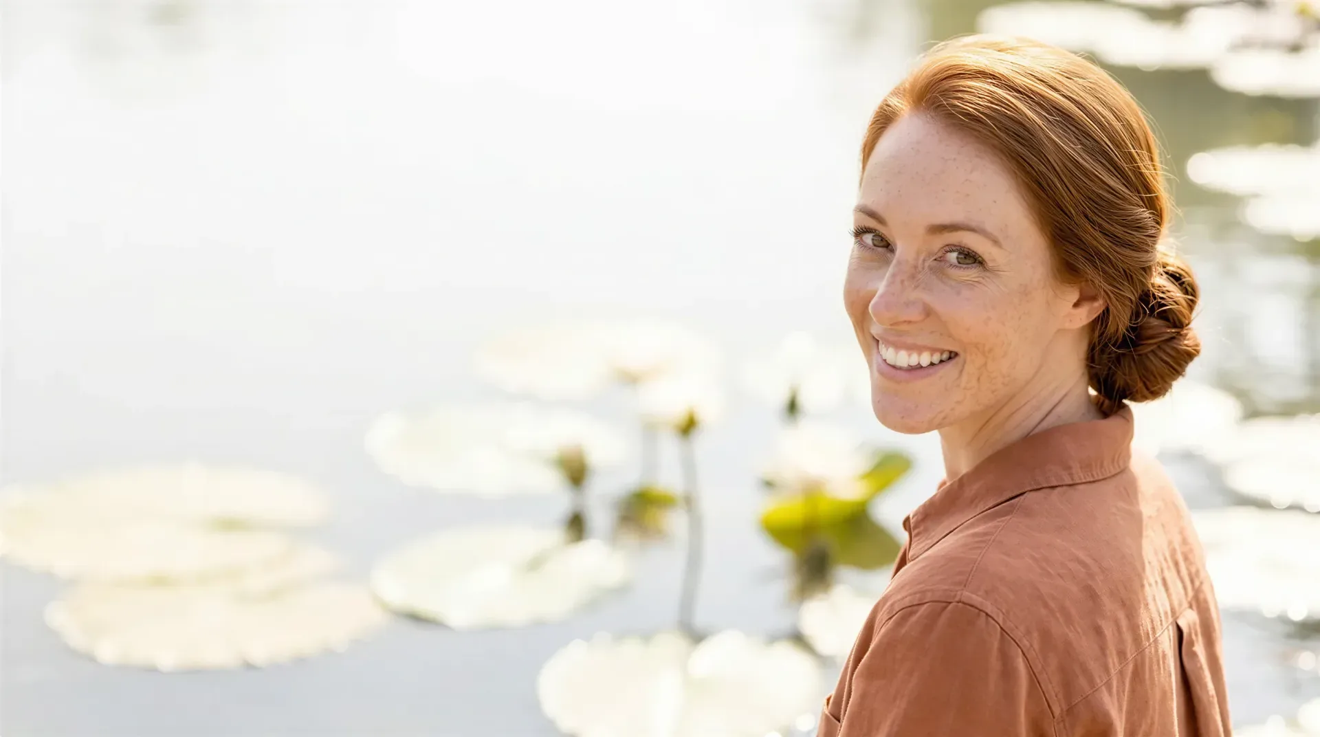 Red-haired woman smiling beside a lily pond in soft daylight near Dallas, reflecting the softer frown lines Jeuveau delivers