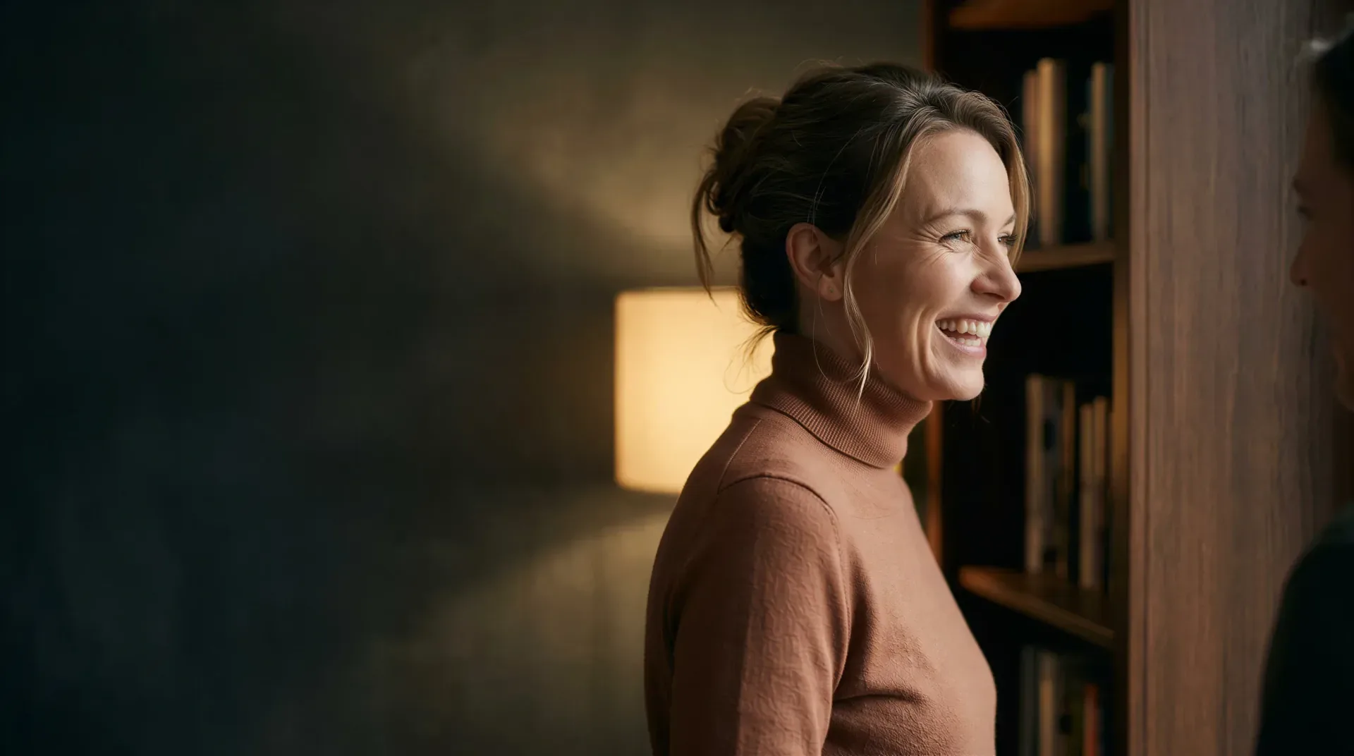 Woman laughing warmly in profile in a softly-lit book-lined room, evoking natural-looking dermal filler and facial balancing care in Dallas