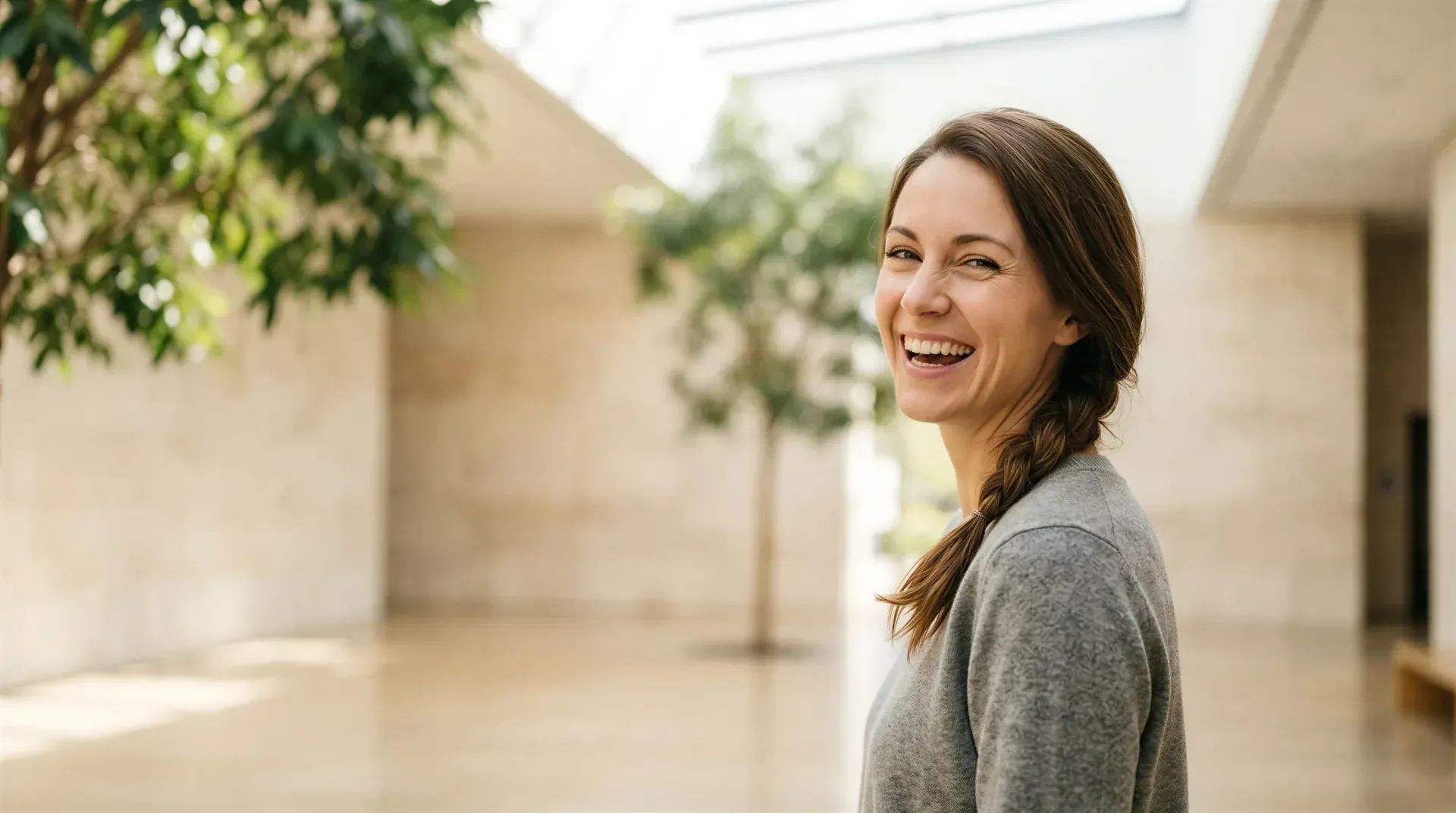 Woman with a side braid laughing in a bright, plant-filled atrium, evoking reassuring laser tattoo and PMU removal care in Dallas