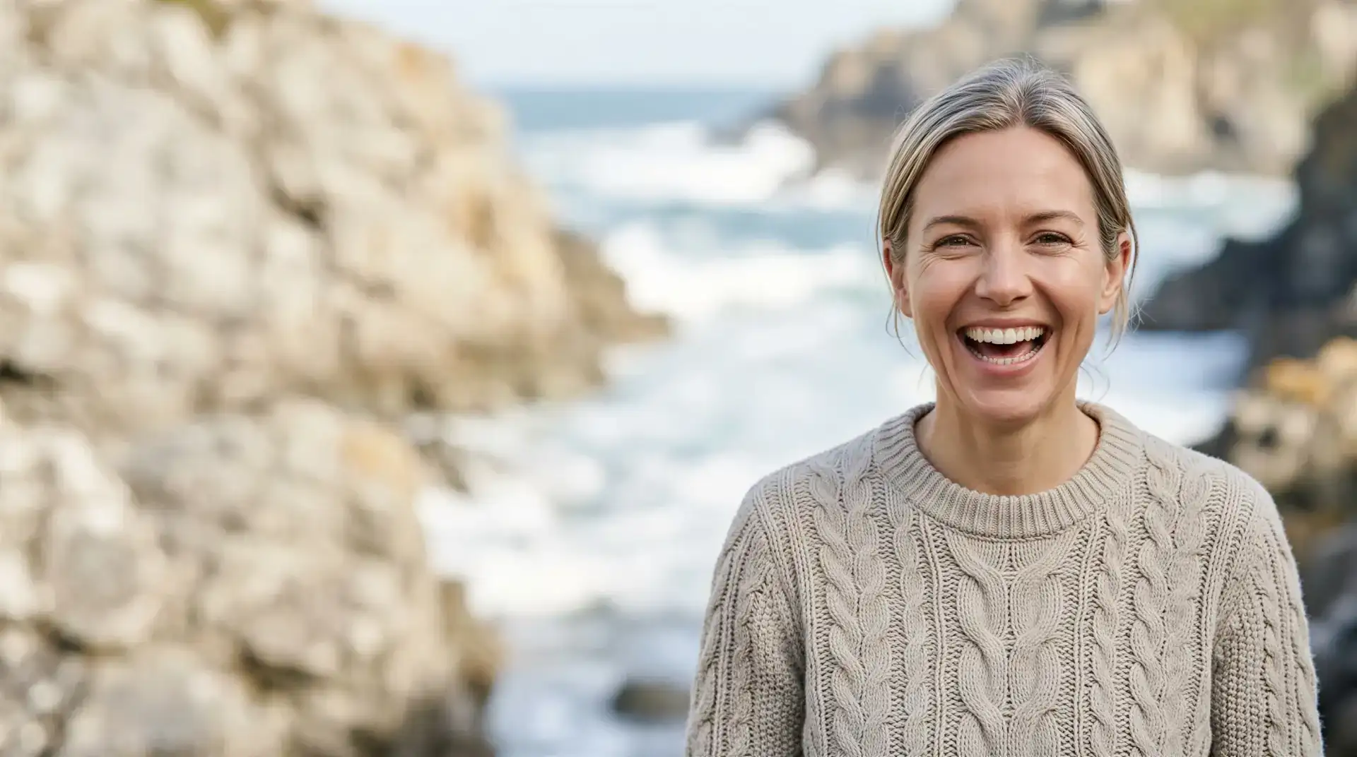 Smiling woman with windswept hair on a sunlit rocky coastline, evoking discreet, confident sexual health and intimacy care in Dallas