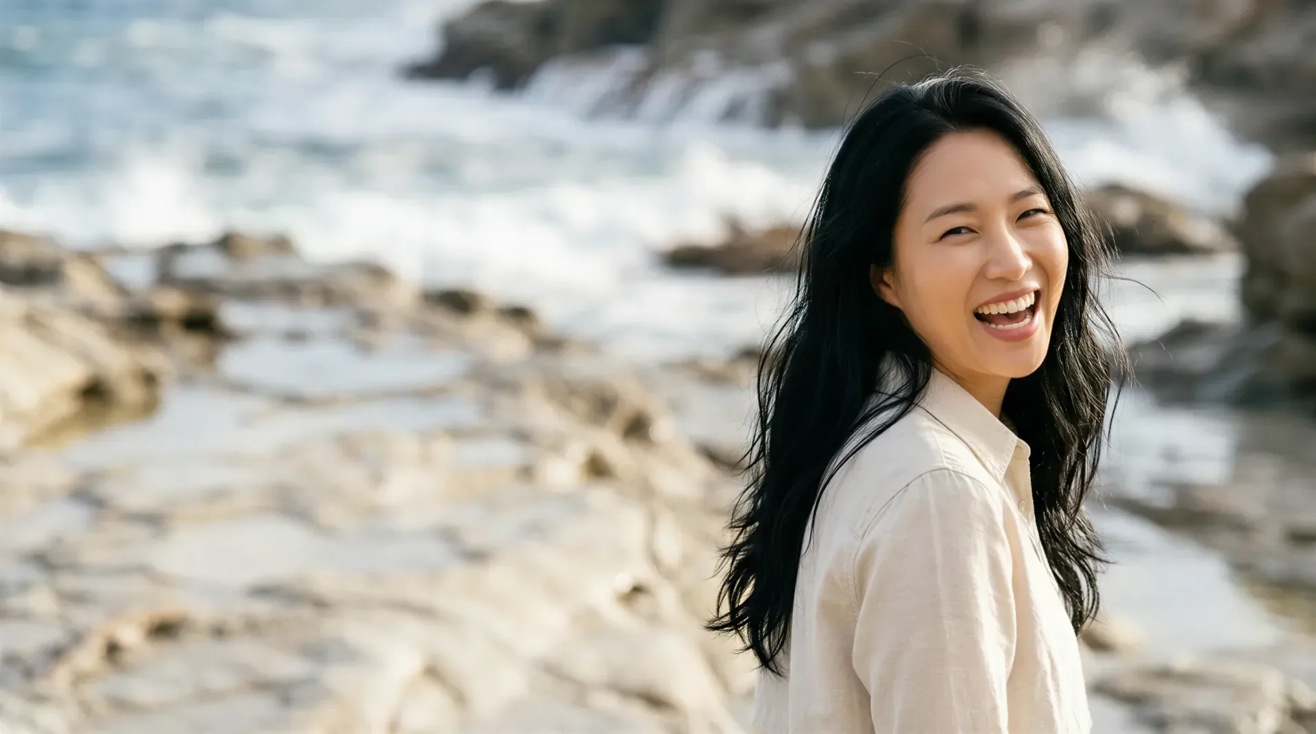 Woman laughing on a sunlit rocky coastline with glowing, healthy skin
