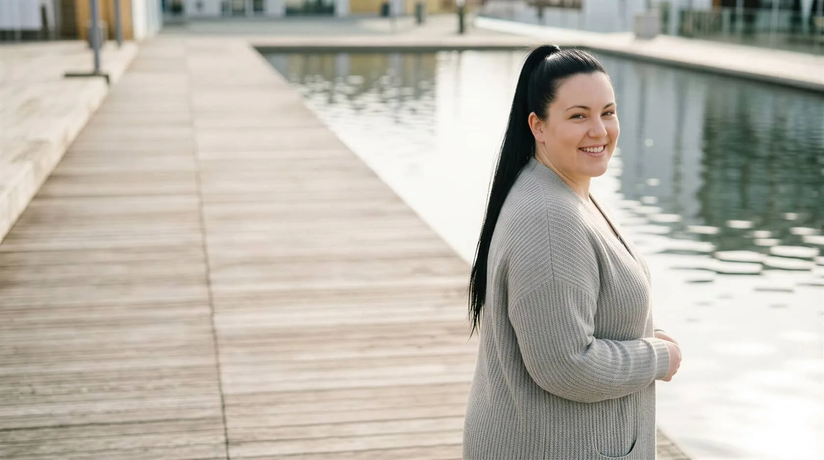 Smiling woman with a dark ponytail beside calm water on a boardwalk, evoking honest permanent makeup removal in Dallas