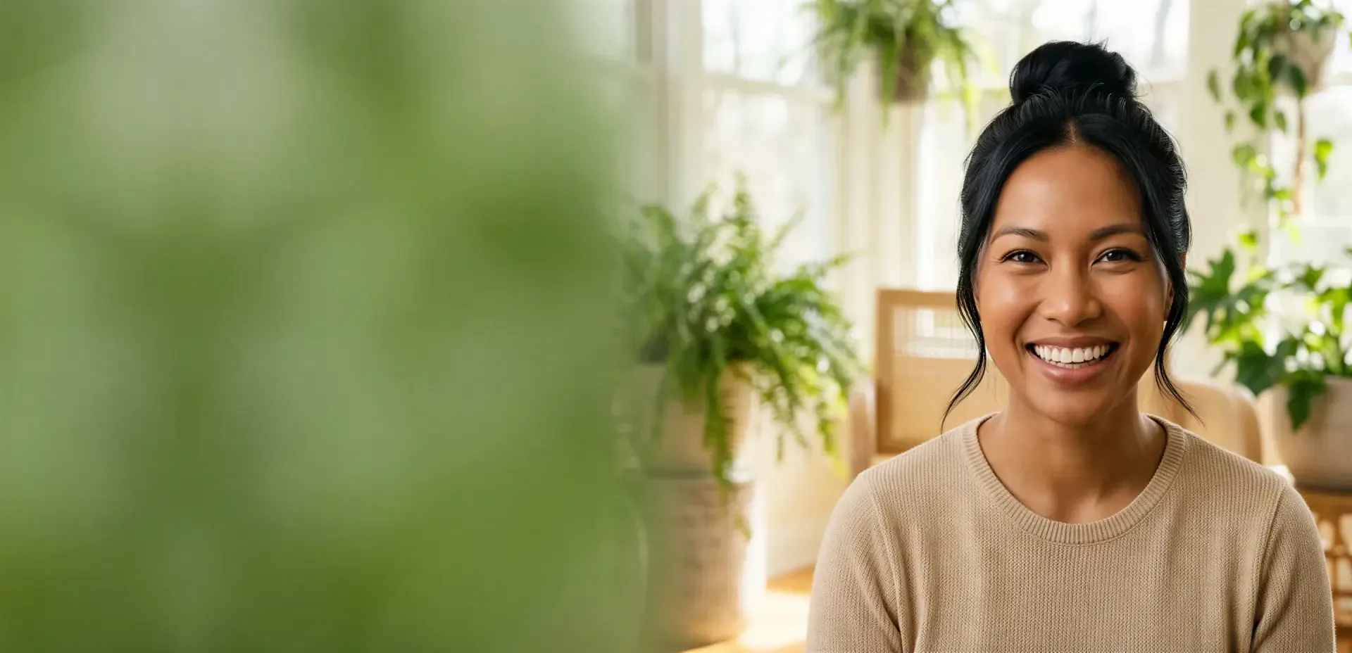 Woman with dark hair in a bun smiling warmly in a bright, plant-filled room, evoking natural Radiesse volume results in Dallas