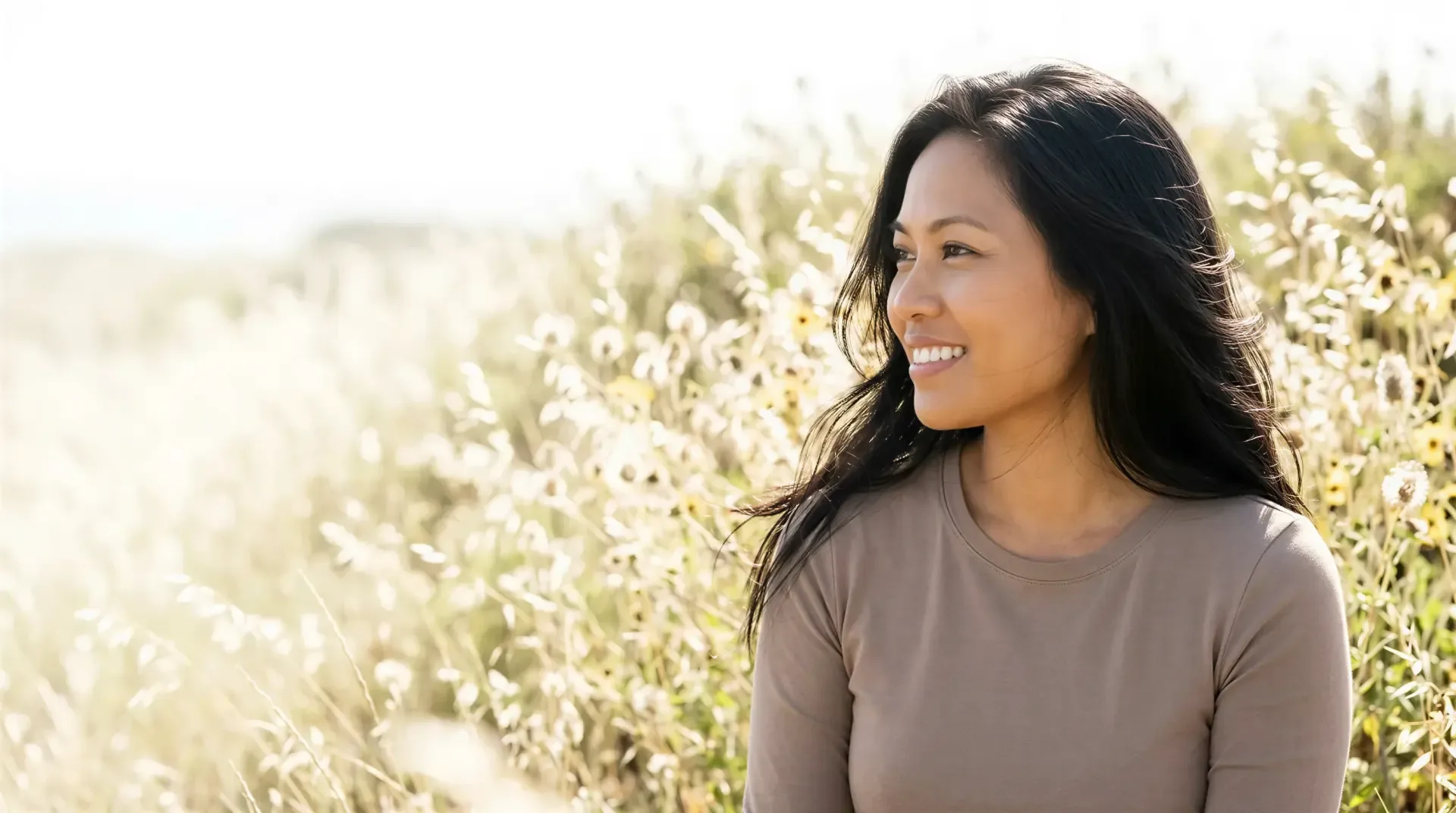 Woman with long dark hair smiling in a sunlit wildflower field, evoking natural-looking RHA filler results in Dallas