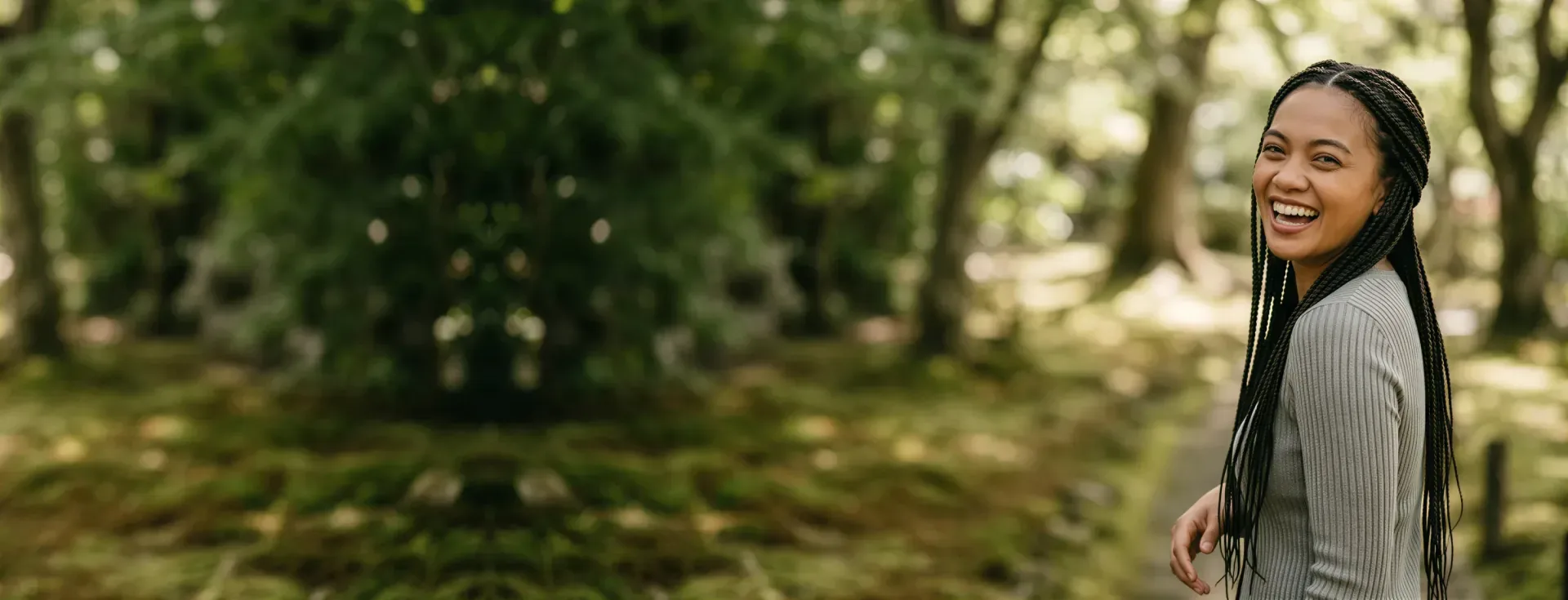 Smiling woman with long braids on a wooded path in dappled light, evoking soft, even Versa filler results in Dallas
