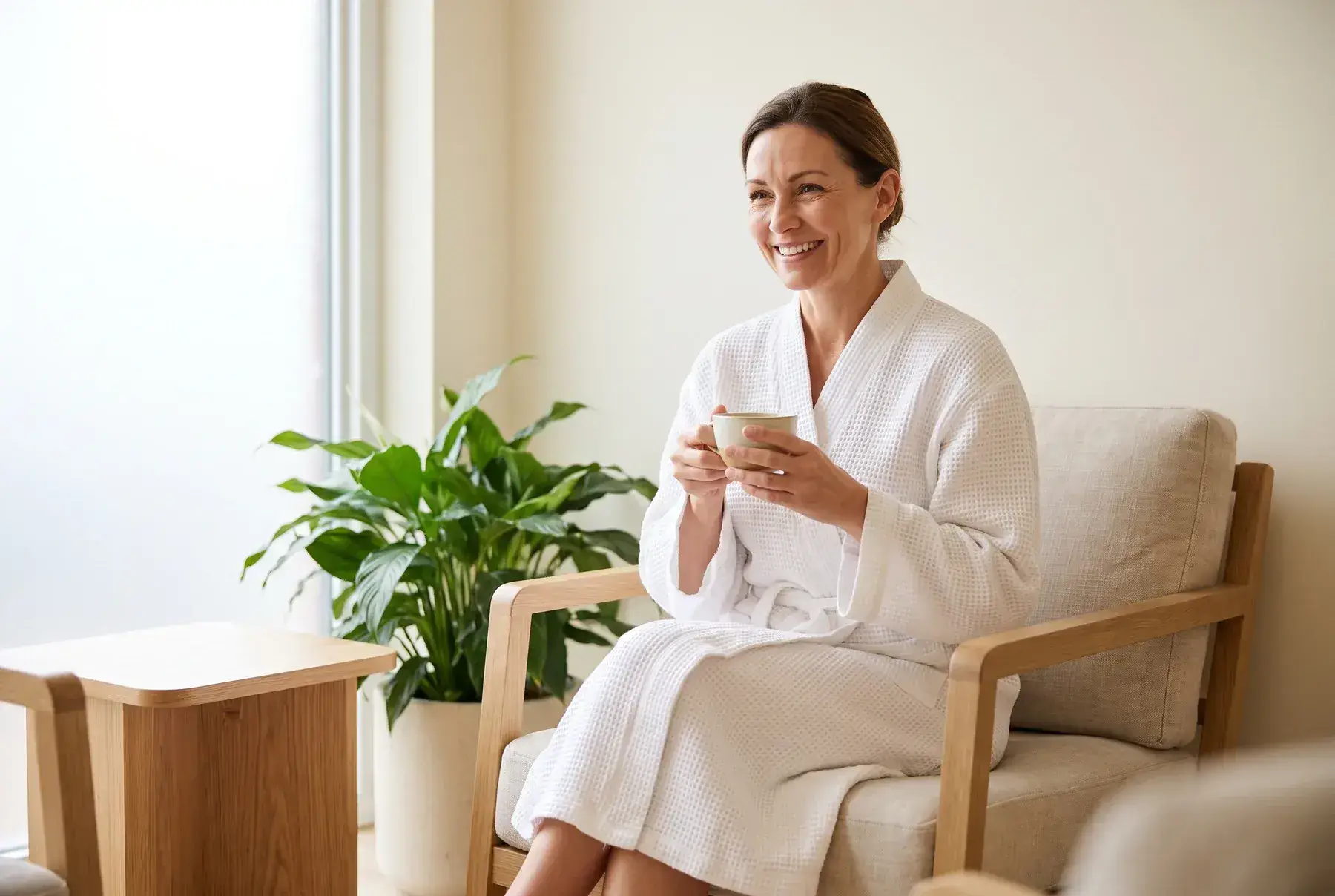 A smiling woman in a white robe enjoying tea in a calm, neutral medical spa lounge, evoking the ongoing care of the Well Labs+ VIP membership