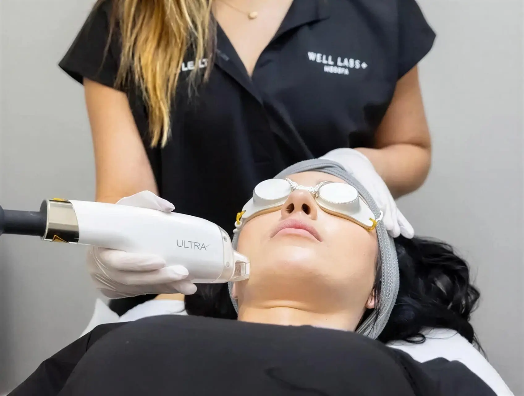 A Well Labs+ provider in white gloves guiding a ULTRA laser handpiece along a client's jaw during a skin resurfacing treatment, with the client wearing protective eye shields