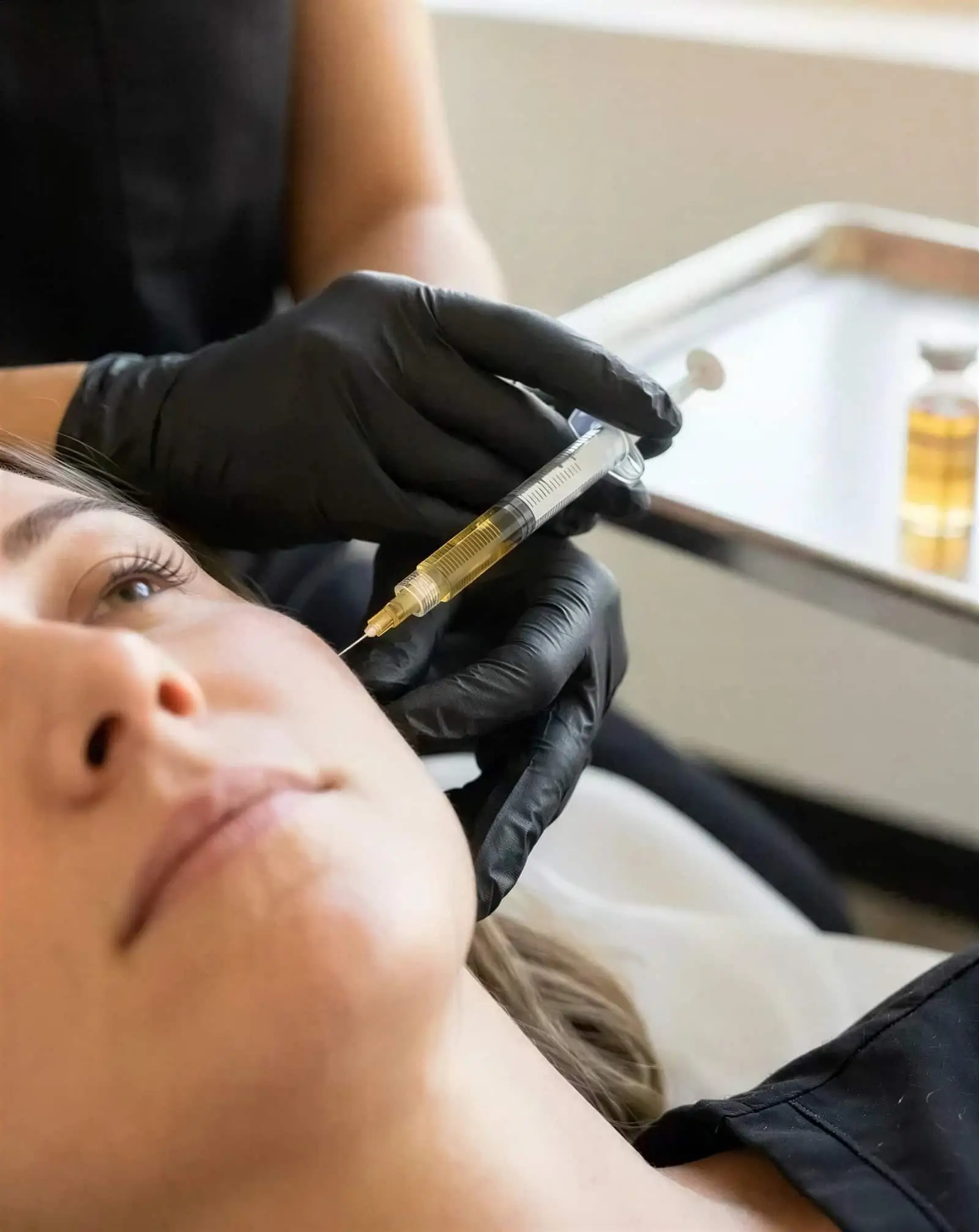 A gloved Well Labs+ provider administering a PRF injection to a reclining client, with golden platelet-rich fibrin on the tray beside her